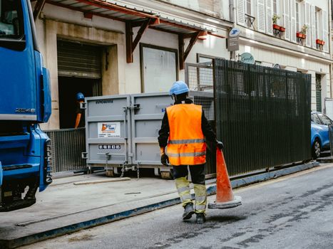 A construction worker in safety gear walks on a city street holding a traffic cone.